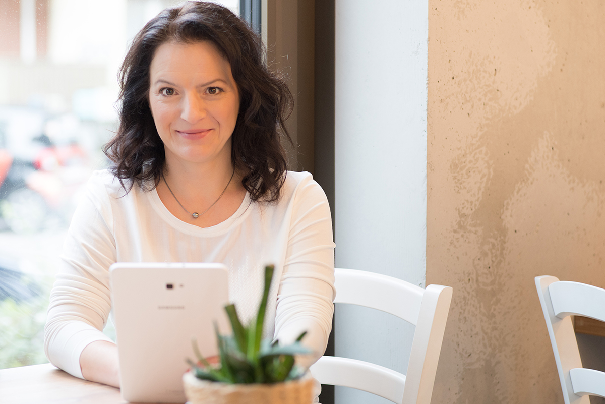 Sandra Brestrich ist Marketingpartnerin. Auf dem Foto sitz sie an einem Tisch und schaut in die Kamera. In der Hand hält sie ein Tablet, auf dem sie offenbar gerade etwas liest.